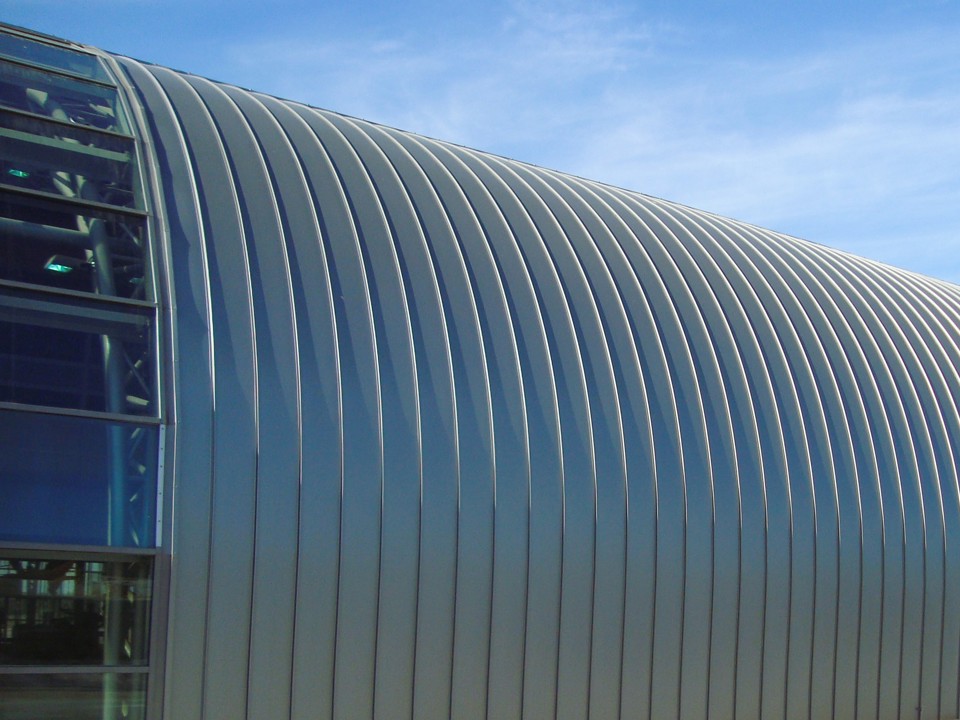 Gebogenes Aluminiumdach einer großen Halle vor einem blauen Himmel.