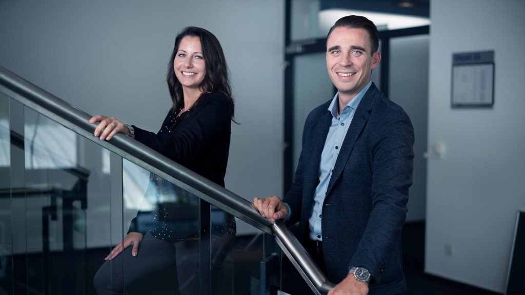 Zwei lächelnde Mitarbeiter in Bürokleidung an einer Treppe im Speira Büro.