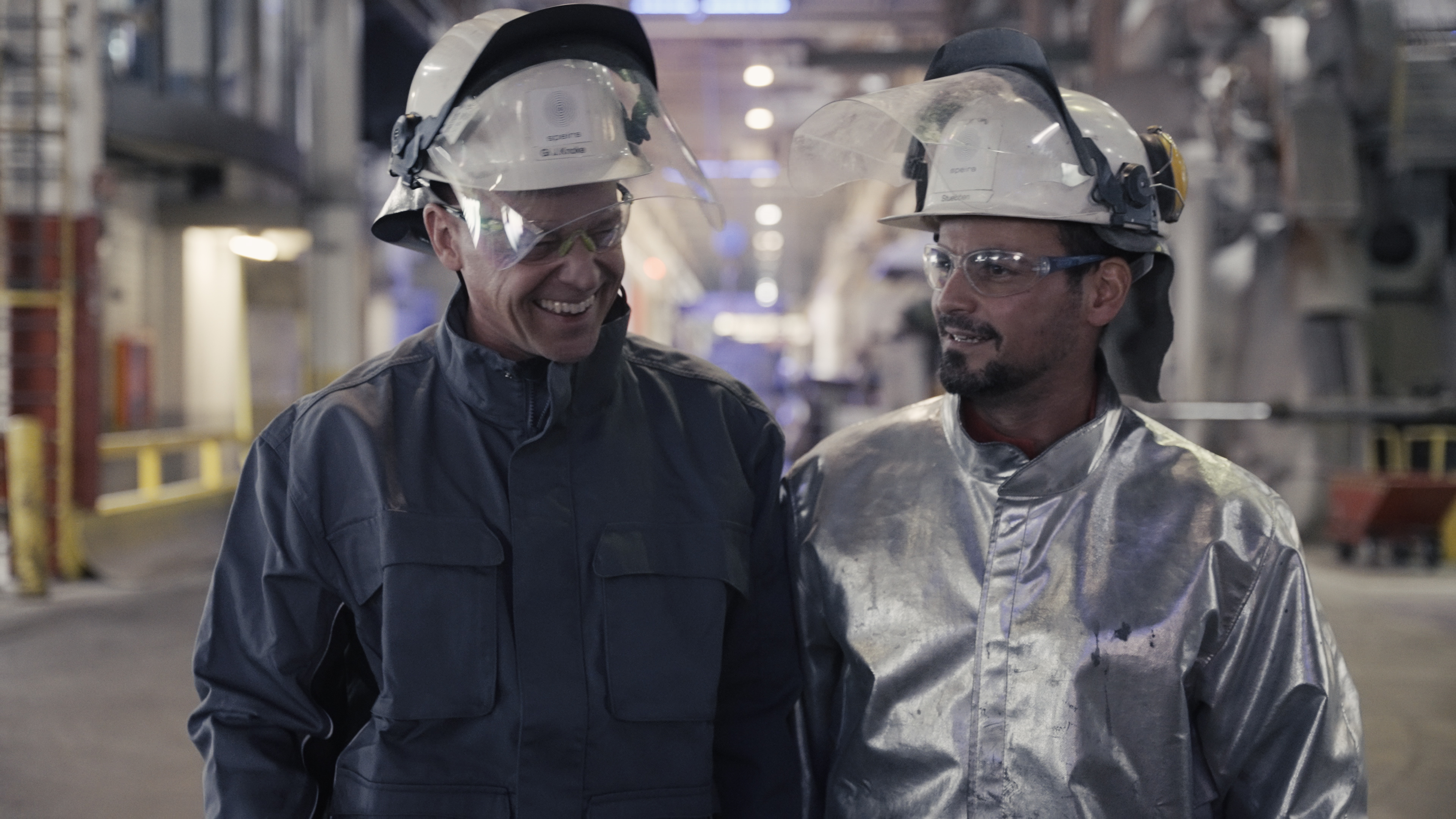 Two smiling Speira employees walk through the aluminum plant in their work clothes.