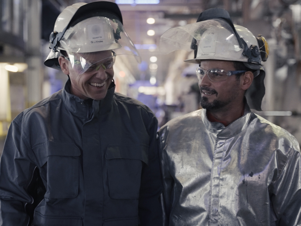 Zwei lächelnde Speira Mitarbeitende laufen in Arbeitskleidung durch Aluminiumwerk.