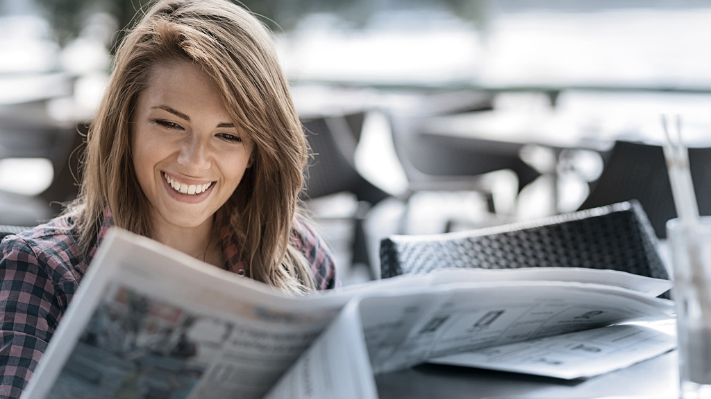 En kvinne sitter utenfor, leser en avis og smiler.