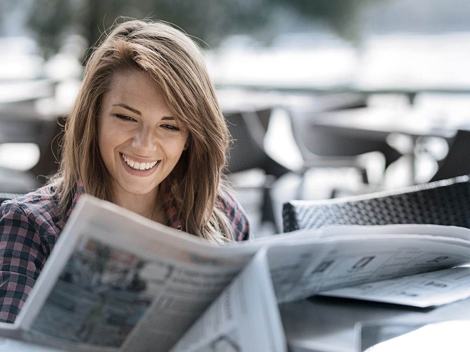 Eine Frau sitzt draußen, liest eine Zeitung und lächelt.