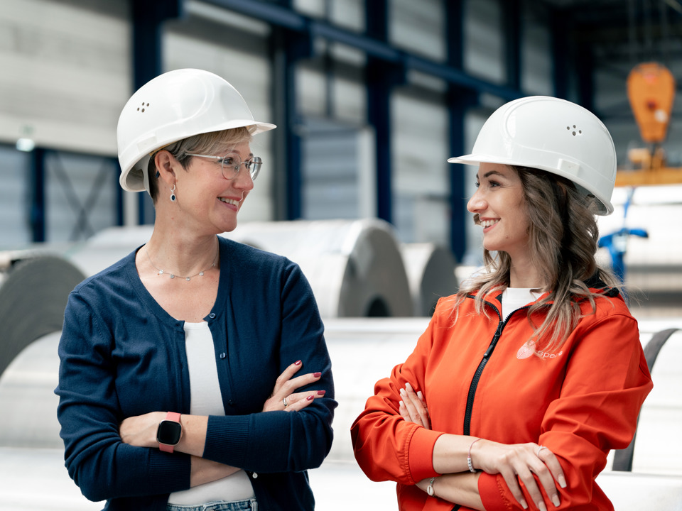 Zwei weibliche Speira-Mitarbeiterinnen in einer Fabrikumgebung lächeln sich an.