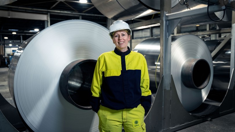 Eine lächelnde Speira Mitarbeiterin in Arbeitskleidung vor gestapelten Aluminium Coils.
