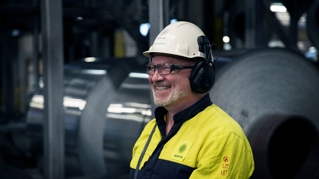 Ein lächelnder Speira Mitarbeiter in Arbeitskleidung vor einem Aluminium Coil.