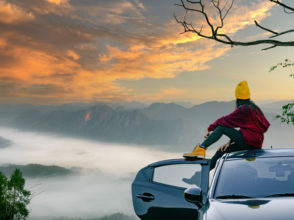 Frau sitzt auf einem Autodach in der Natur mit Blick auf einen Sonnenuntergang.