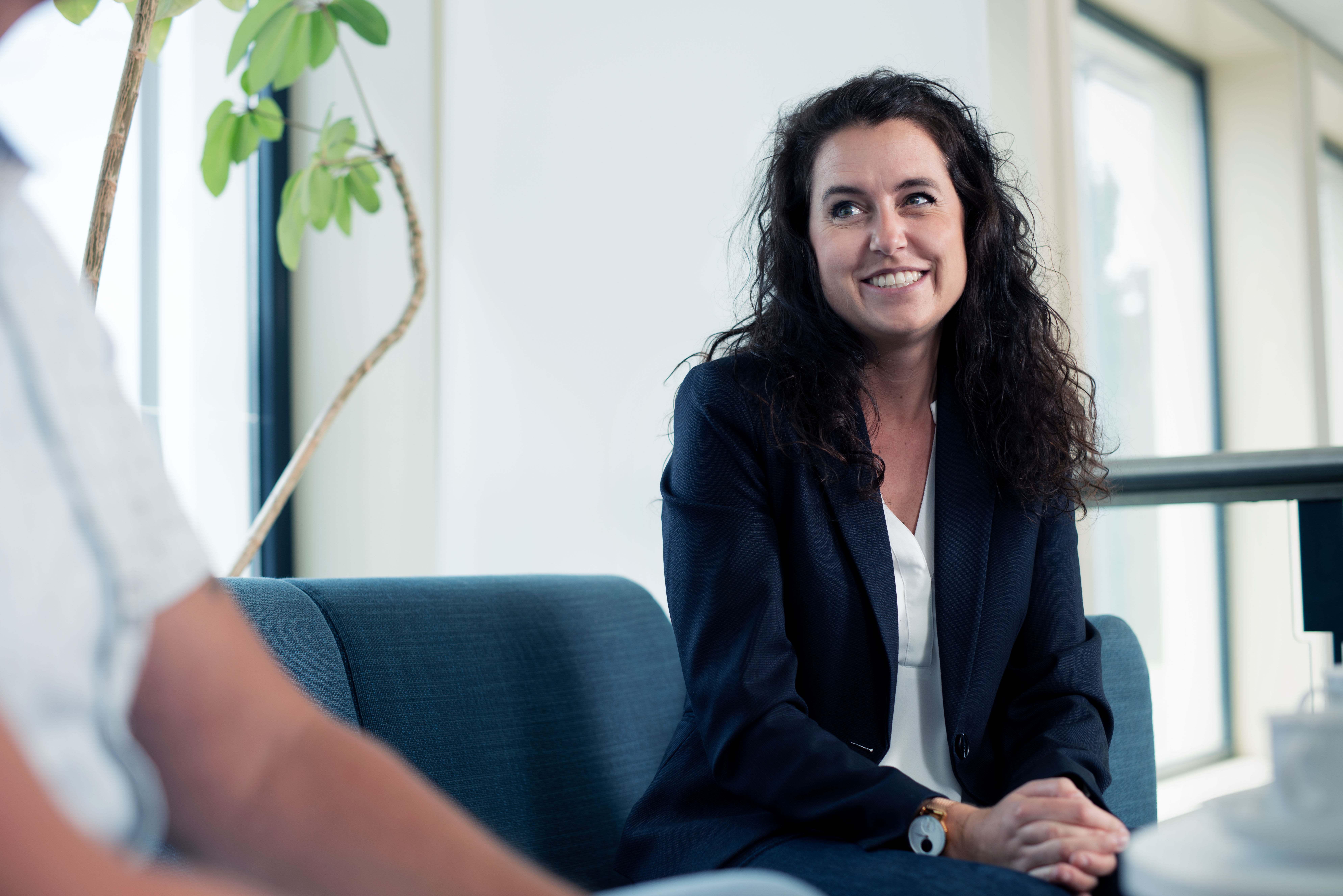 A smiling Speira employee in office attire sits on a sofa in the office.
