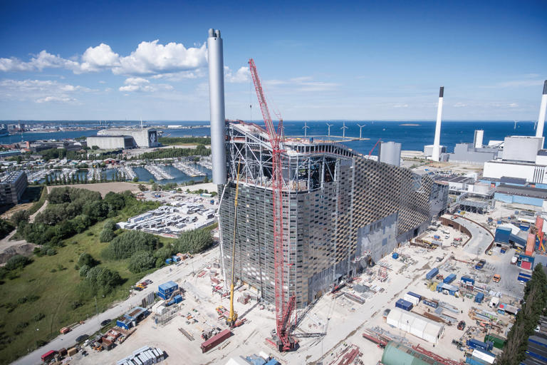 Industribygning med aluminiumsfasade foran et vann og vindturbiner.