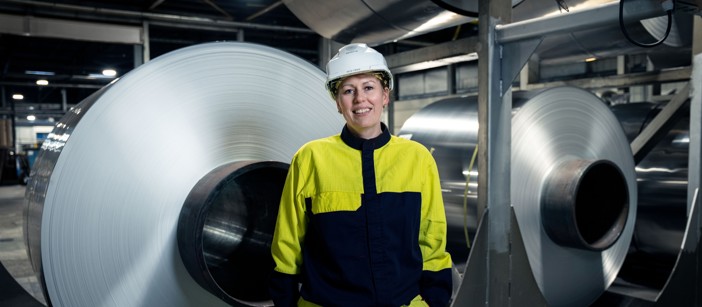 A smiling Speira employee in work clothes in front of stacked aluminum coils.